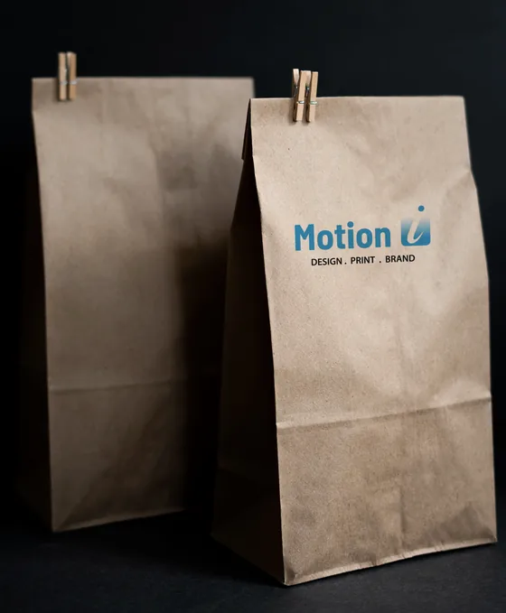 Kraft paper bags on a dark background, one printed with the Motion Imprints logo.